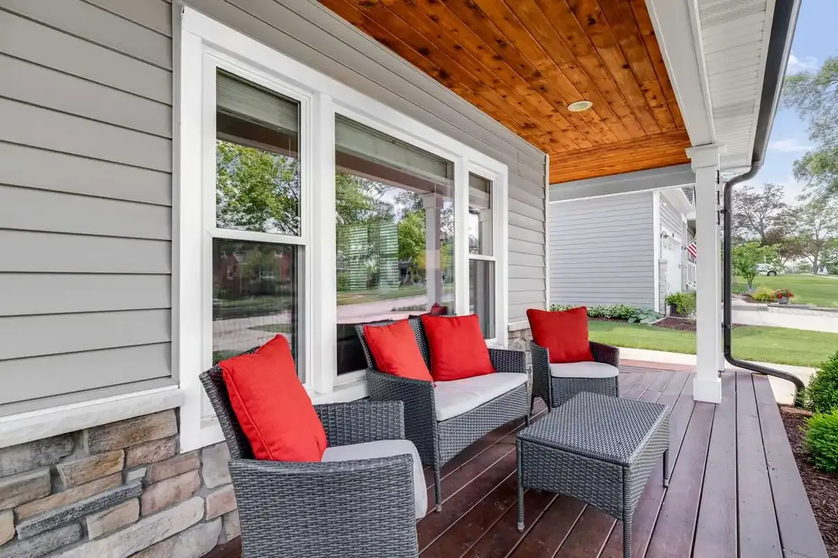 The front porch of a gray house with gray wicker furniture with bright red cushions.